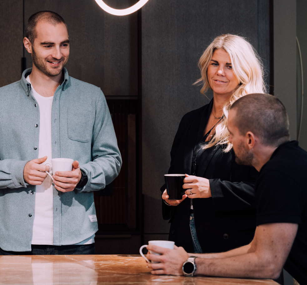 Tre personer står rundt om et bord og holder kaffekopper i Kvass. De ser ud til at være engagerede i en samtale og ser glade ud. En mand har en lysegrå skjorte på, en kvinde med blondt hår har en sort jakke på, og en anden mand har en sort t-shirt på.