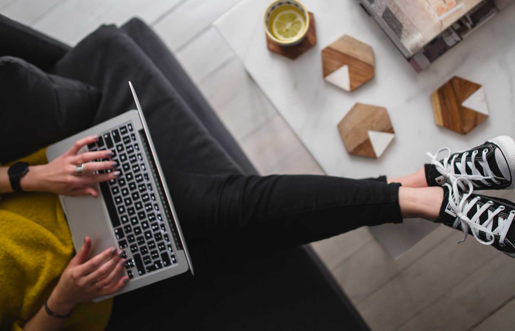 En person sidder afslappet på en sofa med en bærbar computer på skødet. Personen har sorte sneakers på og hviler fødderne på sofabordet, hvor der står en kop med en citronskive. På sofabordet ligger også nogle bøger.